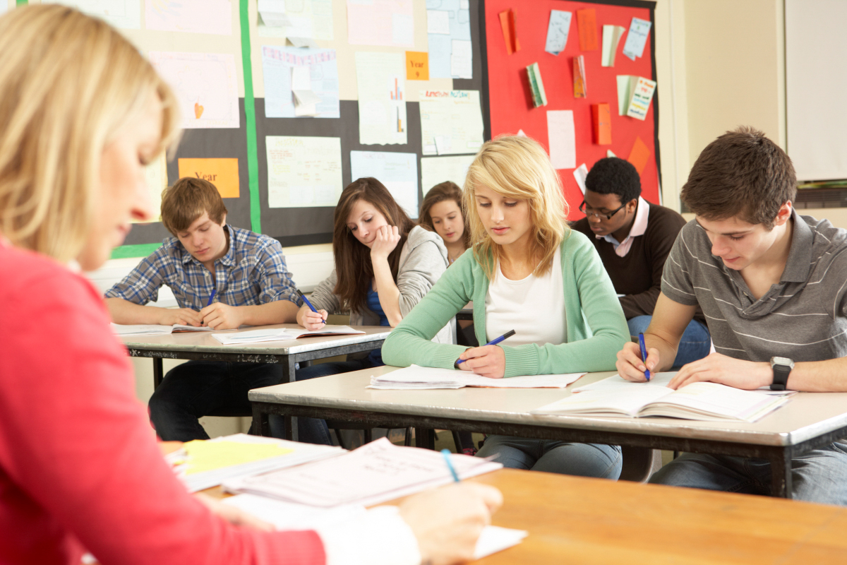 Unterricht im Klassenraum Schülerinnen und Schüler sitzen in einer Klasse und schreiben in Hefte, vor ihnen sitzt eine Lehrerin an einem Pult.