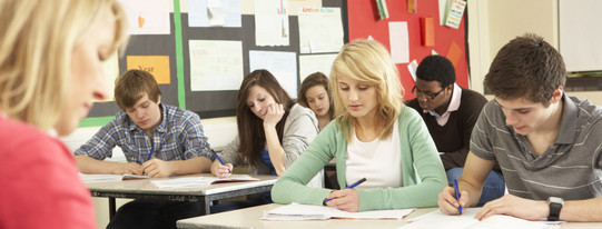 Schülerinnen und Schüler sitzen in einer Klasse und schreiben in Hefte, vor ihnen sitzt eine Lehrerin an einem Pult.