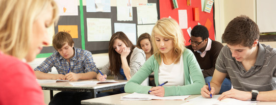 Schülerinnen und Schüler sitzen in einer Klasse und schreiben in Hefte, vor ihnen sitzt eine Lehrerin an einem Pult.