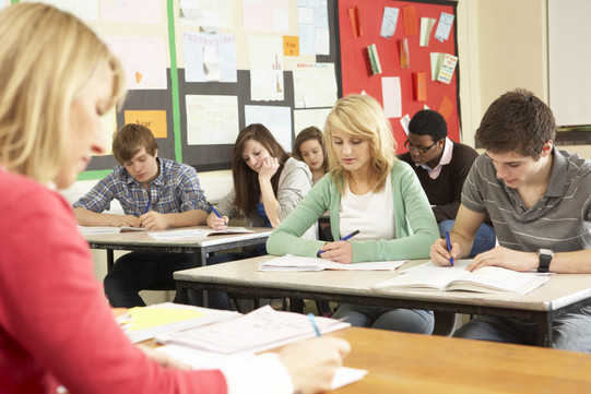 Schülerinnen und Schüler sitzen in einer Klasse und schreiben in Hefte, vor ihnen sitzt eine Lehrerin an einem Pult.