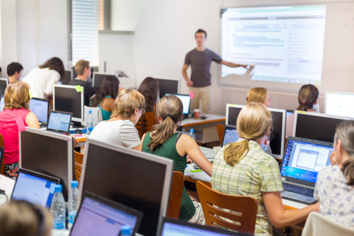 Viele Menschen sitzen in einem Kursraum vor Computern. Vorne steht ein Lehrer und zeigt etwas auf einem Bildschirm.