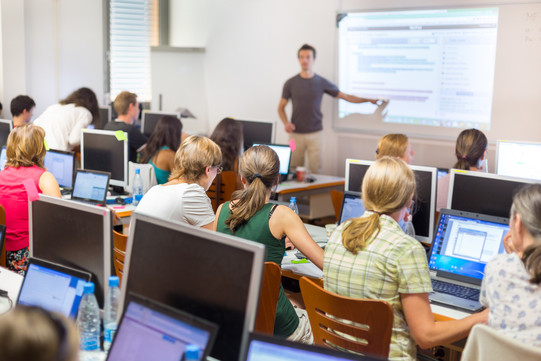 Many people sit in front of computers in a course room and teacher stands in front and shows something on a screen.