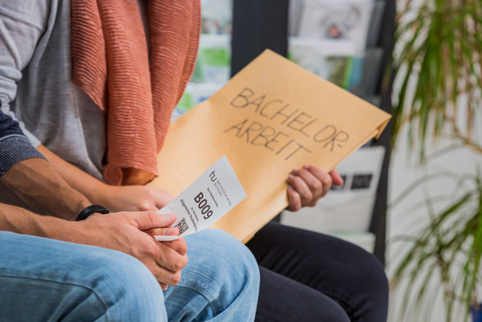 Close-up of two people sitting nect to each other. One is holding an envelope, the other a small piece of paper with a number printed on it